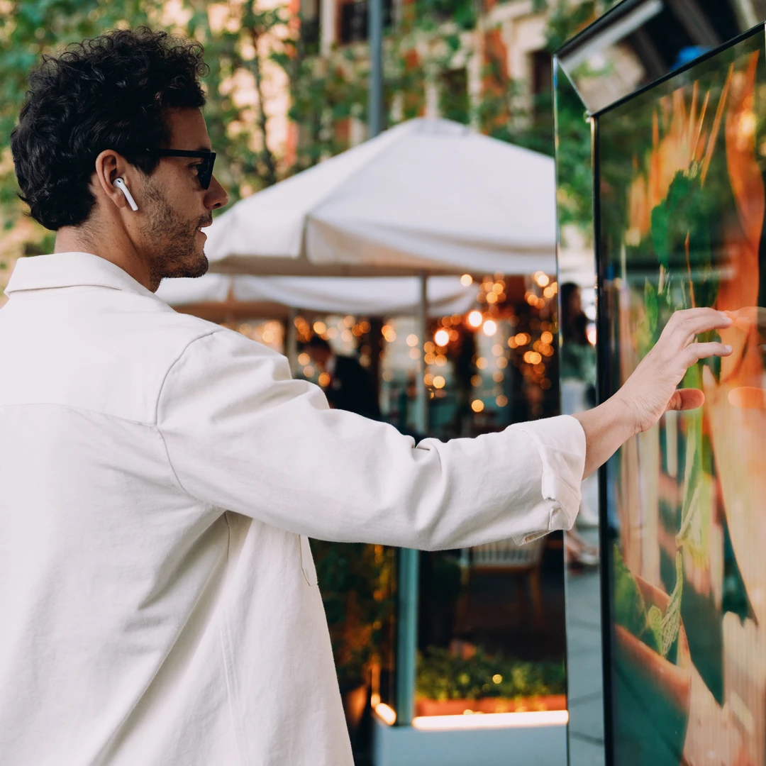 Man in white shirt using outdoor digital kiosk touchscreen with wireless earbuds