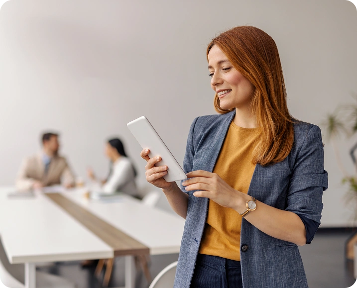 Professional-woman-smiling-while-using-a-digital-tablet-in-a-modern-office-setting-with-two-colleagues-conversing-in-the-background Professional woman smiling while using a digital tablet in a modern office setting, with two colleagues conversing in the background