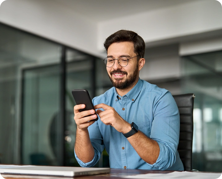 Smiling-professional-man-using-a-smartphone-at-his-office-desk-wearing-glasses-and-a-smartwatch-in-a-modern-workplace-setting Smiling professional man using a smartphone at his office desk, wearing glasses and a smartwatch in a modern workplace setting