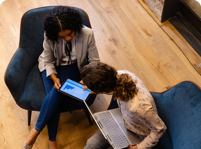 Two businesswomen sitting in modern chairs, collaborating with a tablet and laptop, reviewing data charts in a professional office setting